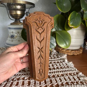 May include: A brown, wooden wall sconce with a carved floral design. The sconce has a rectangular shape with a decorative top and a detailed, symmetrical pattern. The wood grain is visible, and the sconce appears to be vintage.