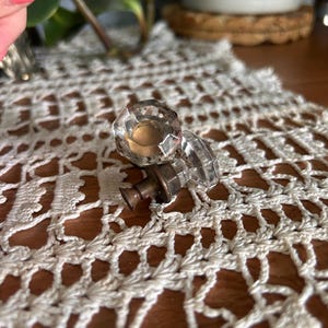 May include: A clear, faceted glass door knob with a metallic, bronze-toned base. The knob features a multi-sided, geometric shape that reflects light, attached to a cylindrical metal stem with a small, flared end piece for mounting purposes.