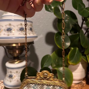 May include: A small, rectangular handbag featuring a white, blue, and gold geometric needlepoint pattern. The bag has an ornate, textured gold-tone metal frame with a decorative clasp and a matching gold-tone metal chain link shoulder strap.