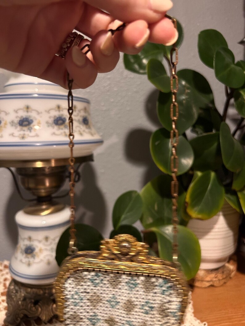 May include: A small, rectangular handbag featuring a white, blue, and gold geometric needlepoint pattern. The bag has an ornate, textured gold-tone metal frame with a decorative clasp and a matching gold-tone metal chain link shoulder strap.