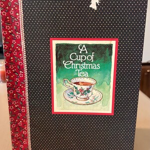 May include: A book titled "A Cup of Christmas Tea" with a black cover featuring white polka dots. The cover has a red border with a floral pattern on the left side. The book's cover art depicts a teacup with tea.