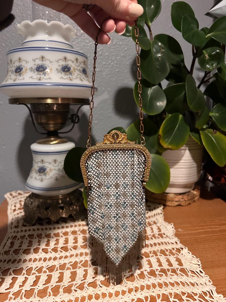 May include: Vintage metal mesh purse with a decorative gold-tone frame and chain handle. The purse features a white, brown, and teal geometric pattern with fringe at the bottom. A classic accessory.