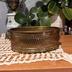May include: A vintage brass planter with a wavy, textured design. The oval-shaped container has small handles on each end. The planter sits on a cream-colored, crocheted doily. A decorative lamp and potted plants are in the background.