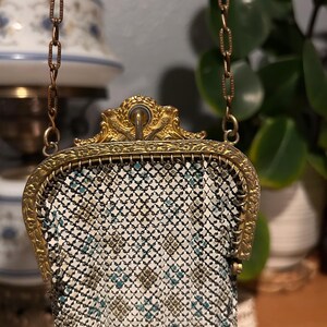 May include: A small metal mesh purse featuring a white, black, and teal geometric pattern. The bag has an ornate gold-tone metal frame with decorative engravings and a matching gold-tone chain strap attached to the top of the frame.