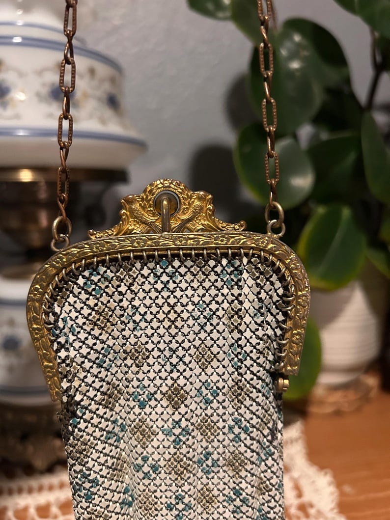 May include: A small metal mesh purse featuring a white, black, and teal geometric pattern. The bag has an ornate gold-tone metal frame with decorative engravings and a matching gold-tone chain strap attached to the top of the frame.