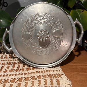 May include: A vintage silver-toned metal serving dish with a lid. The lid features an embossed floral design with apples and a central knob. The dish has two handles and sits on a lace doily.