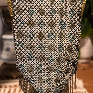 May include: Vintage metal mesh handbag with a geometric pattern in white, black, teal, and gold. The purse has a gold-toned frame and a fringe of metal chain at the bottom. A classic accessory.