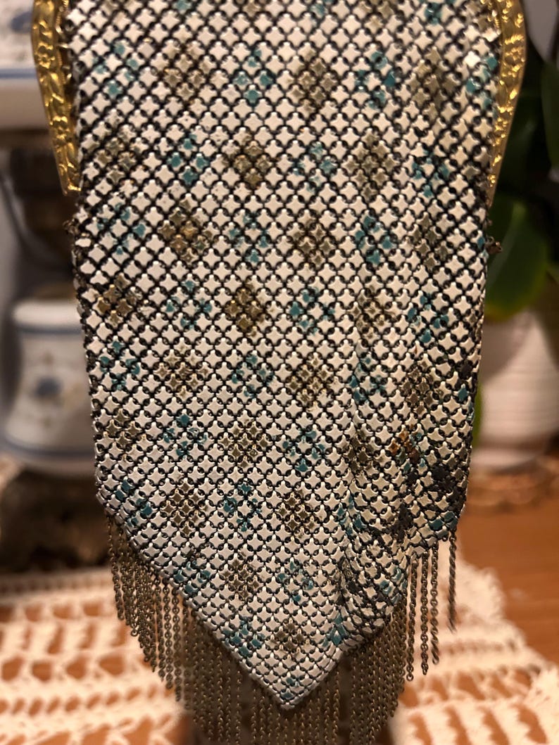 May include: Vintage metal mesh handbag with a geometric pattern in white, black, teal, and gold. The purse has a gold-toned frame and a fringe of metal chain at the bottom. A classic accessory.