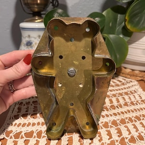 May include: Vintage brass teddy bear cookie cutter. The cutter has a rectangular shape with a bear-shaped cutout in the center. The brass has a tarnished appearance, showing signs of age and use. The cutter is held by a hand with red nail polish.