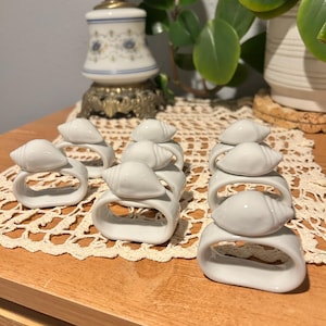 Puede incluir: Un conjunto de servilleteros de cerámica blanca, cada uno con un diseño de concha. Los servilleteros están colocados sobre una superficie de madera con un tapete de encaje. Una lámpara vintage y plantas en macetas se ven en el fondo.