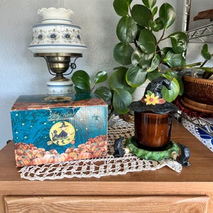 Puede incluir: Una lámpara de aceite vintage con un diseño floral blanco y azul se encuentra junto a una caja con temática de Halloween. También hay un portavelas decorativo con un sombrero de bruja y dos figuras de gatos negros. Una planta verde está en el fondo.
