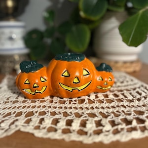 Peut inclure: Un bougeoir en céramique en forme de trois citrouilles orange reliées entre elles avec des tiges vert foncé. Chaque citrouille présente un visage de lanterne sculpté avec des yeux triangulaires et une large bouche peinte en jaune.