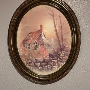May include: Oval framed artwork featuring a quaint cottage scene. The cottage has a red roof and is surrounded by lush greenery and a stone wall. The frame is a decorative gold color, adding a vintage aesthetic to the artwork.