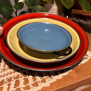 May include: A stack of colorful enamelware dishes. A red plate, a yellow plate, and a blue bowl with a black handle. The plates and bowl have silver rims. The dishes are on a wooden surface.