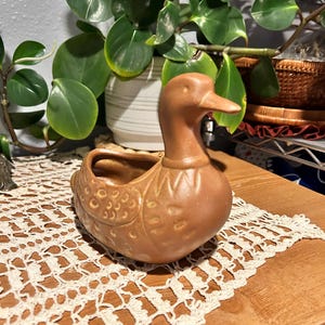 Puede incluir: Una maceta de cerámica marrón con forma de pato y un diseño detallado. El pato tiene un pico largo y una pequeña abertura para plantar. La maceta está sobre una superficie de madera con un tapete de encaje crema.