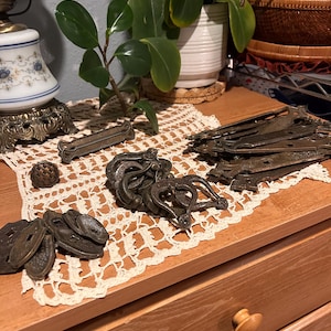 Puede incluir: Una colección de piezas de ferretería metálica antiguas, incluyendo bisagras y tiradores decorativos, dispuestas sobre un tapete de encaje color crema. La ferretería tiene un acabado oscuro y envejecido, lo que sugiere un uso vintage o reutilizado. Una cómoda de madera con un pomo redondo está en primer plano.