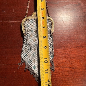May include: An antique metal mesh purse with a gold-colored frame and chain handle. The purse is approximately 7 inches long and features a pattern of white, teal, and brown squares. The bottom edge has a fringe of metal chain.