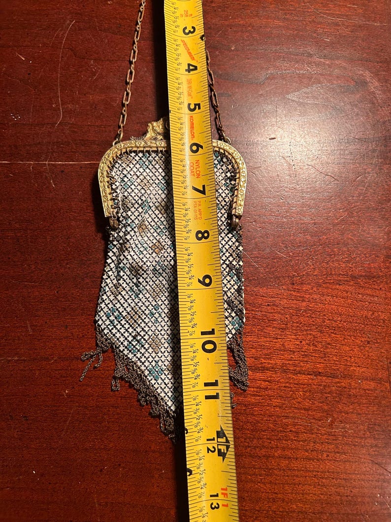 May include: An antique metal mesh purse with a gold-colored frame and chain handle. The purse is approximately 7 inches long and features a pattern of white, teal, and brown squares. The bottom edge has a fringe of metal chain.