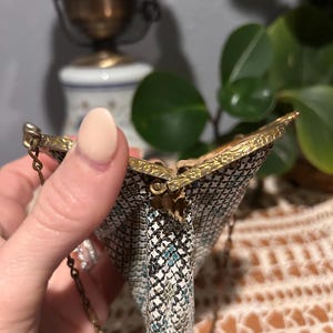 May include: A small metal mesh coin purse featuring a white, black, and teal geometric pattern. The purse has a gold-toned metal frame with an embossed floral design and a thin gold-toned metal chain handle attached to the sides.