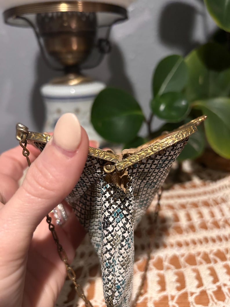 May include: A small metal mesh coin purse featuring a white, black, and teal geometric pattern. The purse has a gold-toned metal frame with an embossed floral design and a thin gold-toned metal chain handle attached to the sides.