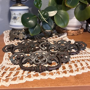 May include: A set of three dark bronze-toned metal drawer pulls featuring ornate, openwork backplates with intricate scrollwork patterns and matching drop handles. Each pull has a central decorative element and a curved, textured handle.