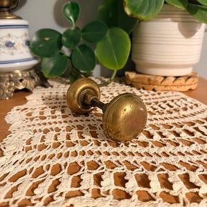 May include: A vintage brass door knob set featuring two round, weathered metallic knobs connected by a central metal spindle. The brass surface shows signs of age, including a dull patina and minor surface imperfections across the hardware.