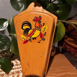 May include: Wooden wall-mounted holder with a rooster illustration. The holder is made of light-coloured wood and features a colourful rooster design with red, yellow, and black accents. The rooster is surrounded by autumn leaves.