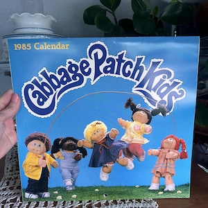 May include: A vintage 1985 Cabbage Patch Kids calendar. The cover features the Cabbage Patch Kids dolls in various outfits against a blue sky background. The calendar is held by a person.