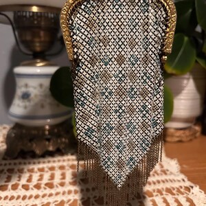 May include: An antique metal mesh purse with a gold-toned frame and chain. The purse features a geometric pattern in white, teal, and gold, with a fringe of gold chains at the bottom. The purse is likely from the early 20th century.