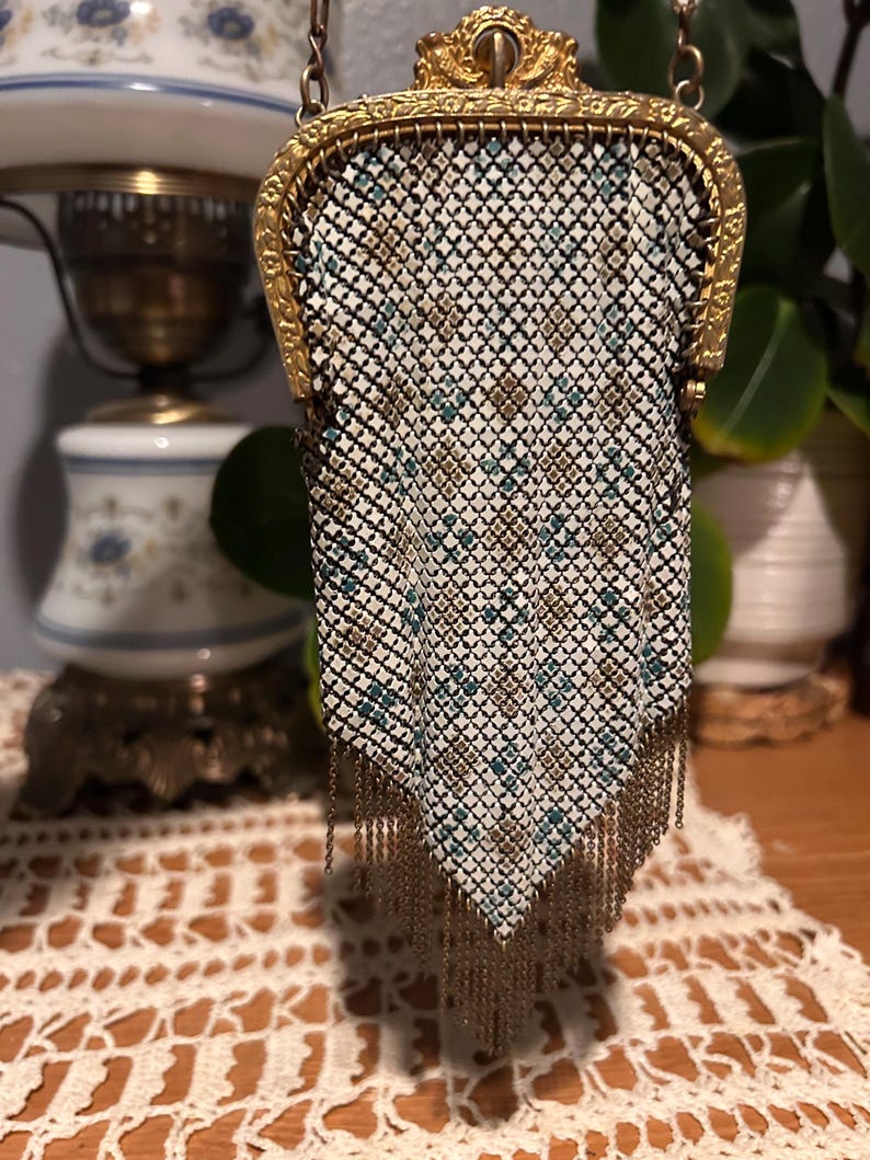 May include: An antique metal mesh purse with a gold-toned frame and chain. The purse features a geometric pattern in white, teal, and gold, with a fringe of gold chains at the bottom. The purse is likely from the early 20th century.