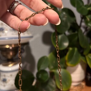 May include: A vintage gold-toned necklace with a delicate chain. The chain is held by a hand, showcasing the intricate link design. The necklace features a decorative pendant with a central stone. The background includes greenery and antique decor.