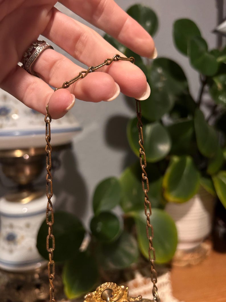 May include: A vintage gold-toned necklace with a delicate chain. The chain is held by a hand, showcasing the intricate link design. The necklace features a decorative pendant with a central stone. The background includes greenery and antique decor.