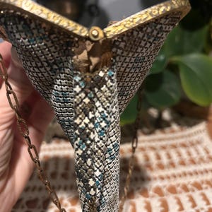 May include: Vintage metal mesh purse with a gold-toned frame and chain strap. The purse features a geometric pattern in shades of silver, teal, and black. The clasp is a small, round gold-toned button. The purse is a classic accessory.