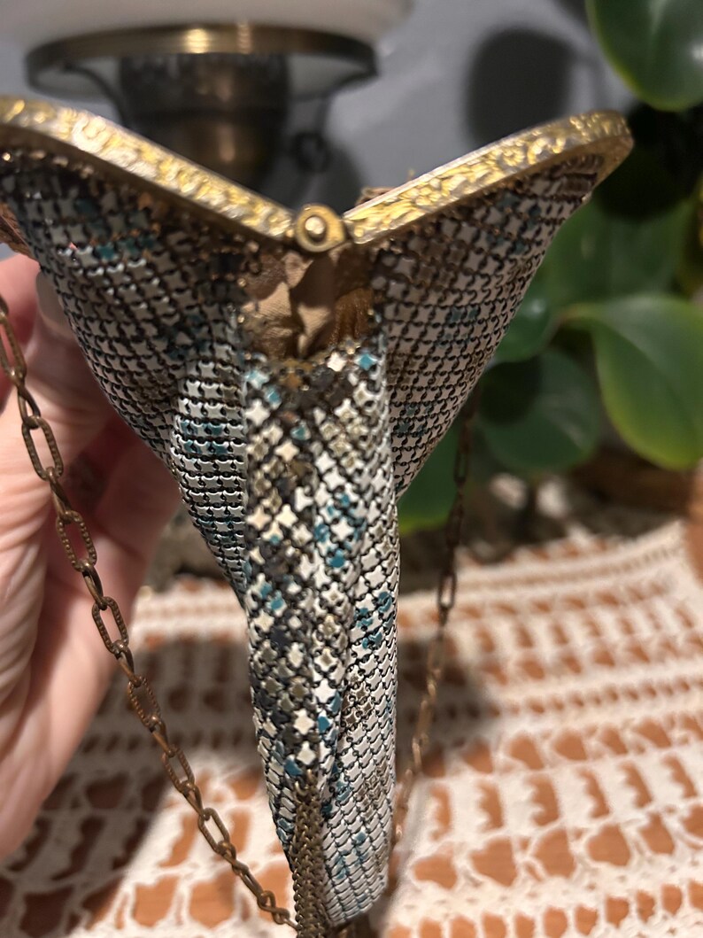 May include: Vintage metal mesh purse with a gold-toned frame and chain strap. The purse features a geometric pattern in shades of silver, teal, and black. The clasp is a small, round gold-toned button. The purse is a classic accessory.