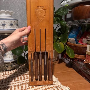 May include: A vintage wooden knife block with five knives. The block is light brown with a carved emblem that reads "Flint". The knives have wooden handles and stainless steel blades. The knife block is on a wooden table.