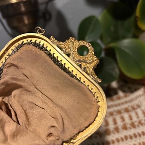 May include: A gold-toned metal frame purse featuring an ornate, embossed decorative clasp with a circular cutout. The interior is lined with a light tan fabric, and the exterior edge shows a fine, dark metallic mesh construction.