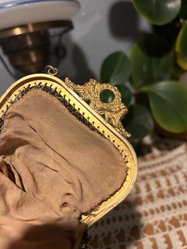 May include: A gold-toned metal frame purse featuring an ornate, embossed decorative clasp with a circular cutout. The interior is lined with a light tan fabric, and the exterior edge shows a fine, dark metallic mesh construction.