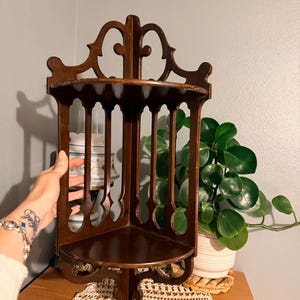 May include: A dark brown wooden corner shelf featuring two triangular tiers connected by vertical slats with arched cutouts. The top piece has a decorative, curved crest design. The structure is designed to fit into a corner, providing two flat surfaces for display.