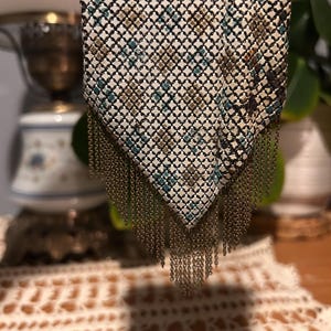 May include: A metal mesh handbag featuring a pointed bottom edge with hanging chain fringe. The surface has a white base with a repeating geometric pattern in shades of teal, gold, and dark brown, creating a textured, grid-like appearance.