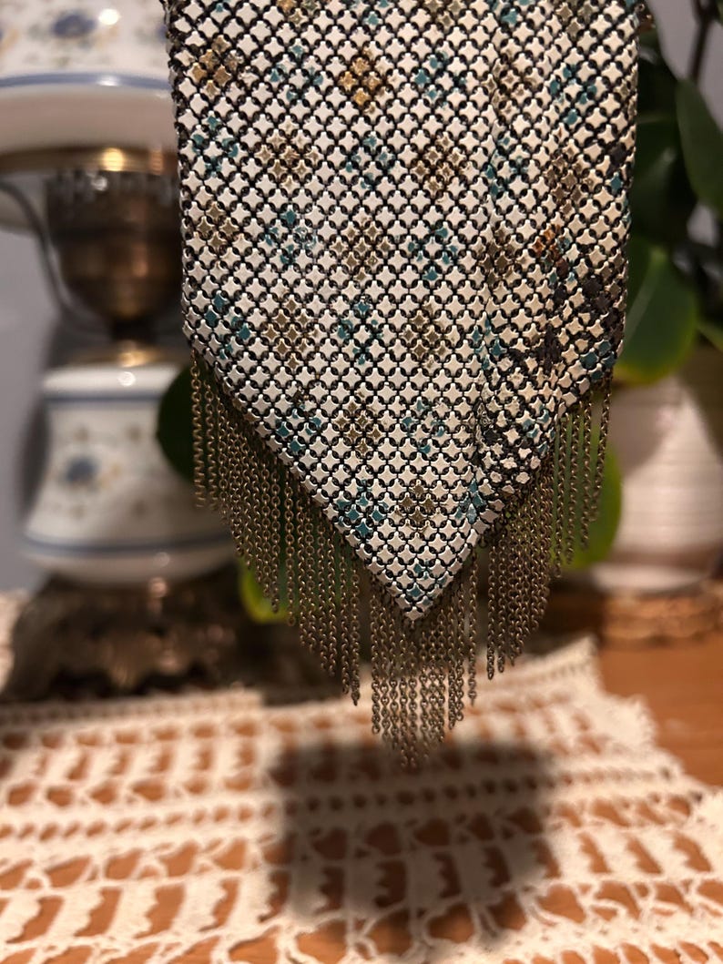 May include: A metal mesh handbag featuring a pointed bottom edge with hanging chain fringe. The surface has a white base with a repeating geometric pattern in shades of teal, gold, and dark brown, creating a textured, grid-like appearance.