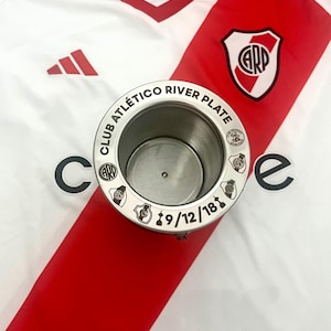 Könnte beinhalten: Ein silberner Metallbecher mit den Worten "Club Atlético River Plate" und dem Datum "19/12/18" eingraviert am Rand. Der Becher steht auf einem weißen und roten Fußballtrikot mit dem River Plate Logo.