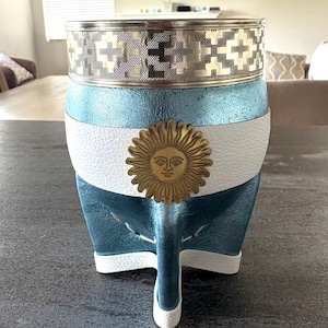 Può includere: Un contenitore per mate con bordo in argento metallizzato dal motivo geometrico, corpo rivestito in pelle testurizzata azzurra e bianca e un emblema solare dorato centrale. Il recipiente è sostenuto da tre piedini con inserti in pelle bianca.