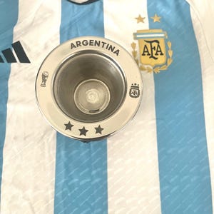 Puede incluir: Una copa de metal plateado con la palabra "ARGENTINA" grabada alrededor del borde. La copa tiene tres estrellas en la parte inferior y una pequeña imagen de un balón de fútbol y las letras "AFA" en el lateral.