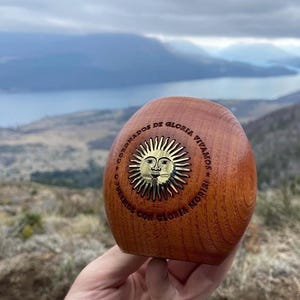 Op de afbeelding: Een houten, eivormig object met een gouden zonnesymbool. Het symbool toont een gezicht en de tekst "CORONADOS DE GLORIA VIVAMOS CON GLORIA MORIRI". Het object heeft een warme bruine kleur en lijkt van gepolijst hout te zijn.