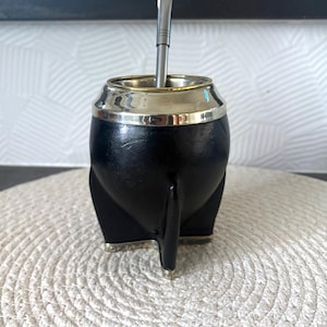 Può includere: Un contenitore per mate nero con un corpo liscio e arrotondato e tre piedi appuntiti alla base. Il bordo superiore è rifinito con una fascia in metallo color argento lucido e una lunga e sottile cannuccia bombilla in metallo è inserita al centro.