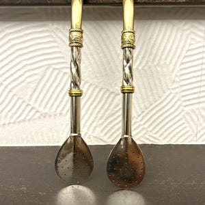 Può includere: Due cannucce bombilla in metallo argentato per tè mate, ciascuna dotata di un filtro piatto perforato a forma di cucchiaio alla base, uno stelo intrecciato e fasce decorative dorate con intricati motivi incisi in alto e al centro.