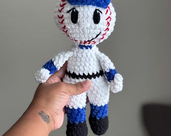 May include: A handmade crochet baseball doll with a smiling face, wearing a blue cap and baseball uniform. The doll is white with red stitching, black accents, and blue details. A fun, whimsical toy.