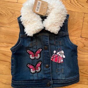 Peut inclure: Un gilet en jean bleu foncé avec un col en fausse fourrure blanche. Le gilet présente deux écussons papillon roses et blancs et un écusson sac à main rose. Le gilet a un devant boutonné et une étiquette attachée.