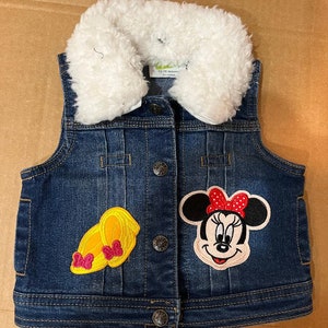 Peut inclure: Un gilet en jean bleu avec un col en fausse fourrure blanche. Le gilet a deux patchs brodés, l'un de Minnie Mouse et l'autre de chaussures jaunes avec des nœuds rouges. Le gilet porte une étiquette indiquant "12-18 mois".