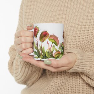 May include: White ceramic mug with a colorful illustration of Venus flytraps. The plants are depicted with red, green, and white details.
